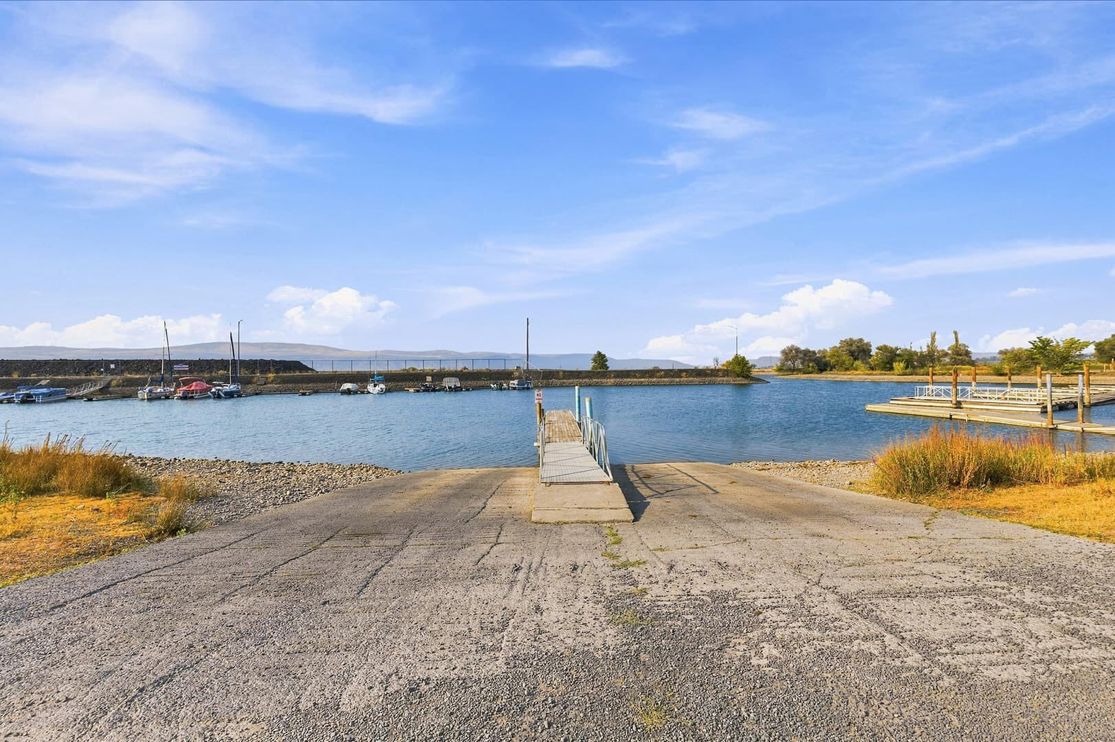 Open shoreline access area with a narrow dock extending into the lake and low hills across the water.