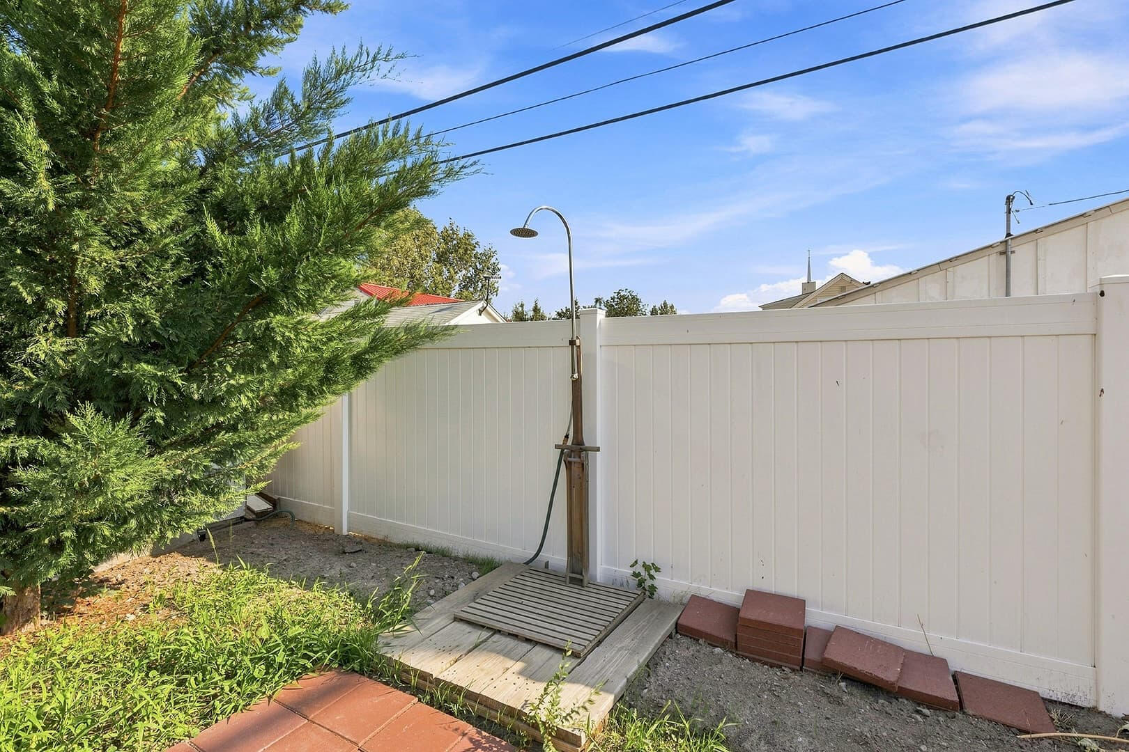 Freestanding outdoor shower beside a white privacy fence, with a small platform and paving below.