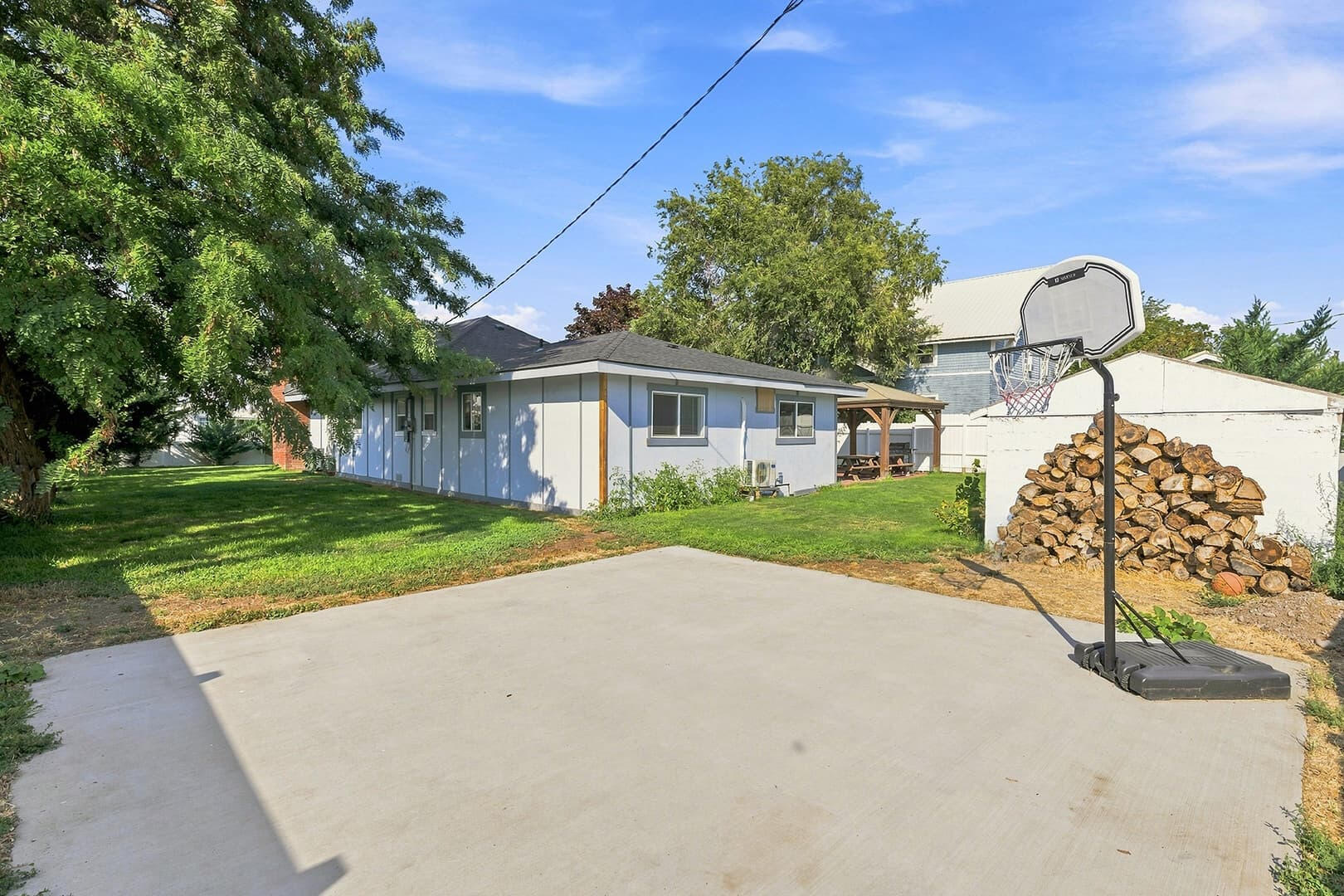 Paved outdoor area with a basketball hoop, with the lawn, gazebo and home beyond.