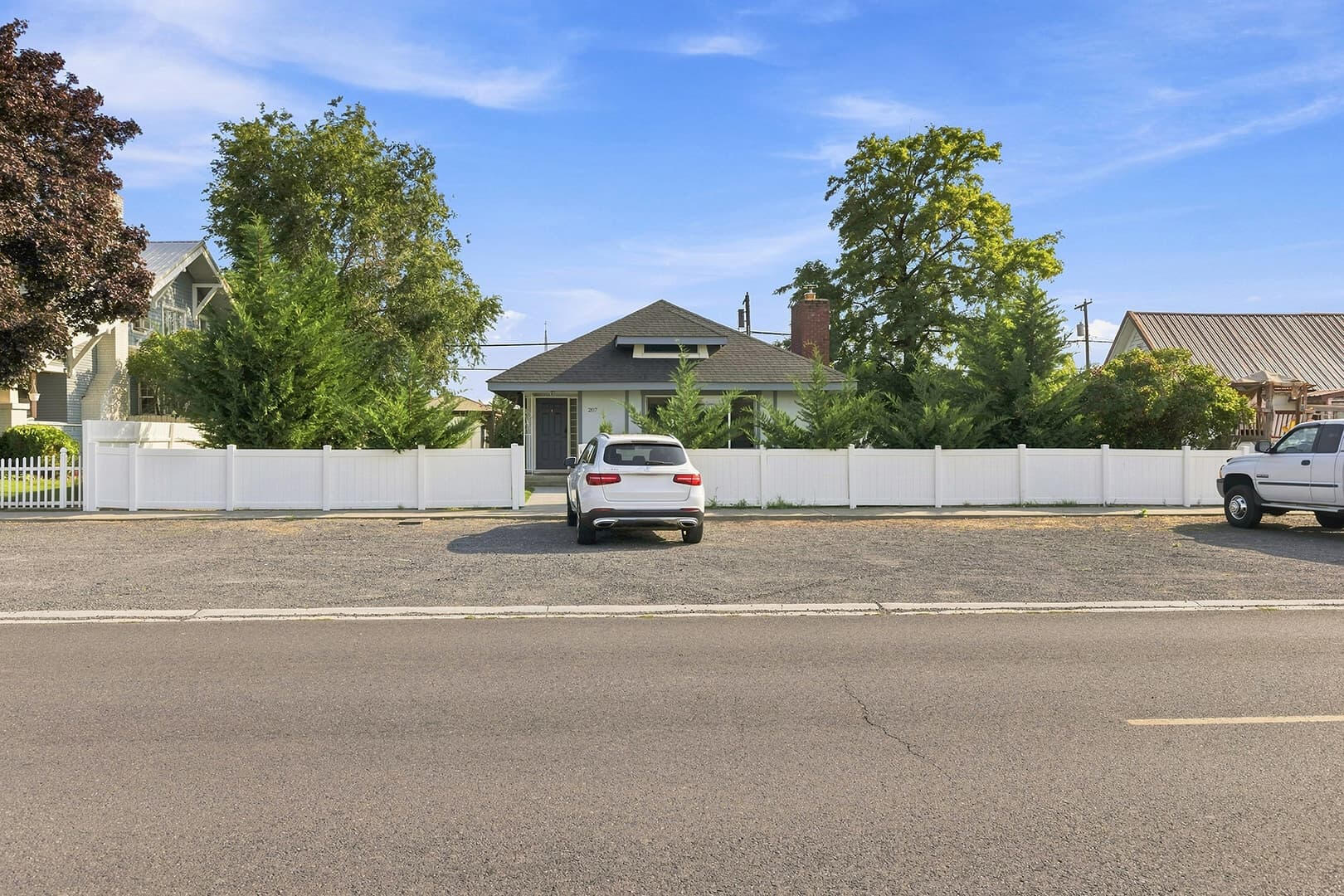 Gravel parking area along the front fence, with parked vehicles and the house behind the trees.