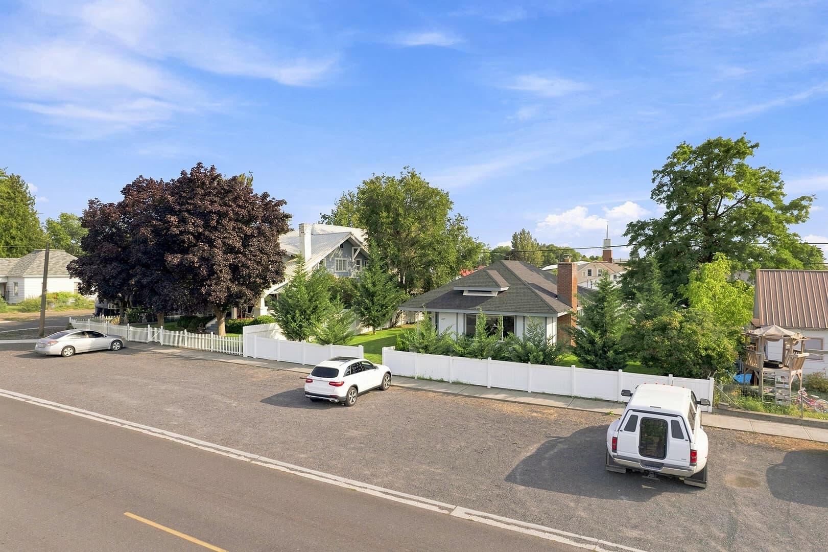 Elevated view of the house, front lawn and white fence, with parked vehicles along the street.