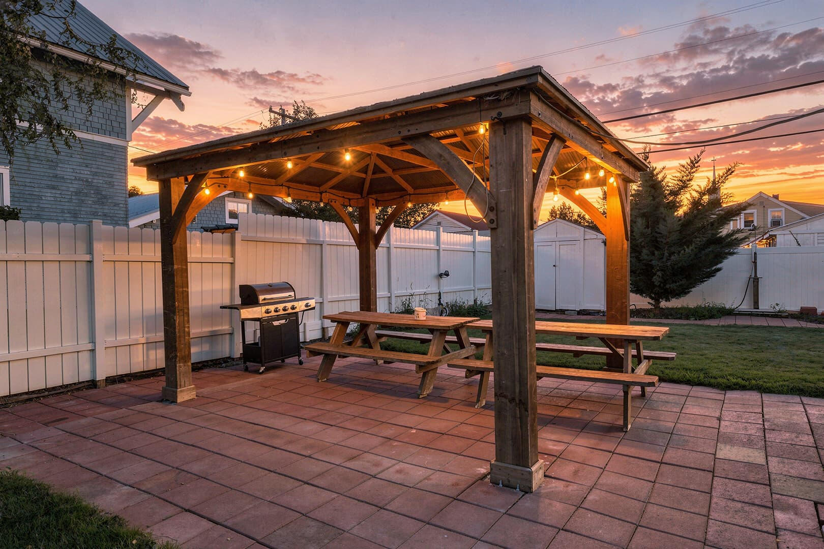 Wooden gazebo over a picnic table on the patio at twilight, with warm lights and a colorful sky.